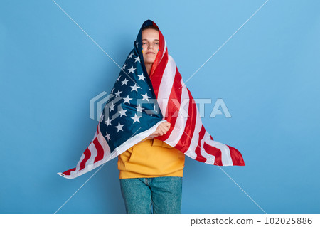 Portrait of proud woman with flag of Ukraine on her cheek, standing wrapped in american flag, looking at camera, feels pride, support from America, posing isolated over blue background. 102025886