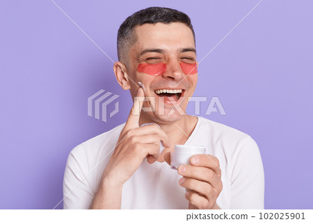 Image of happy man with patches under eyes laughing and keeping finger on cheek, doing morning procedures, expressing happiness, posing isolated on purple background. Image of happy man with patches under eyes laughing and keeping finger on cheek, doing morning procedures, expressing happiness, posing isolated on purple background. 102025901