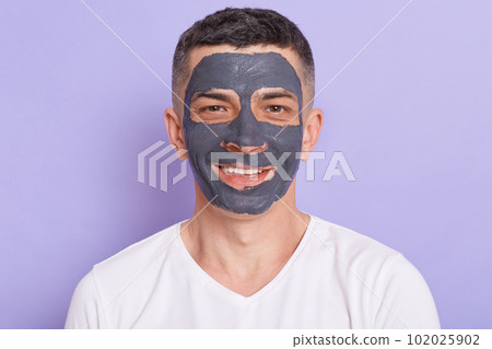 Indoor shot of happy smiling man with clay mask on his face, receiving spa treatments, doing cosmetic procedures, looking at camera with positive facial expression. 102025902