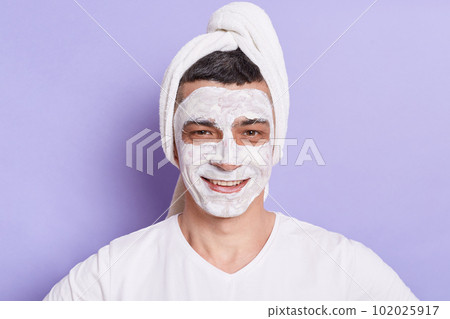 Portrait of smiling Caucasian man with mask for skin, standing wrapped in towel isolated over violet background, doing cosmetology procedures at home. 102025917