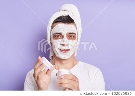 Horizontal shot of well groomed smiling satisfied man with mask for skin doing procedures for moisturizing face, wrapped towel on the head isolated over violet background. Horizontal shot of well groomed smiling satisfied man with mask for skin doing procedures for moisturizing face, wrapped towel on the head isolated over violet background. 102025925