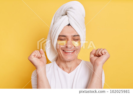 Indoor shot of positive satisfied man wearing white t shirt and towel, having cosmetic patches under his eyes, clenched fists, being extremely happy, posing isolated over yellow background. 102025981