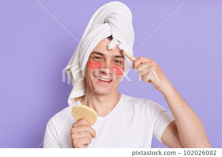 Horizontal shot of smiling handsome man doing cosmetic procedures in morning, standing with patches under eyes and making massage with roller, looking at camera with joyful expression. Horizontal shot of smiling handsome man doing cosmetic procedures in morning, standing with patches under eyes and making massage with roller, looking at camera with joyful expression. 102026082