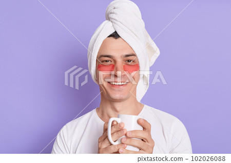 Indoor shot of smiling satisfied man in white towel on head and patches under eyes, holding cup of coffee, enjoying beverage while doing morning cosmetic procedures. 102026088