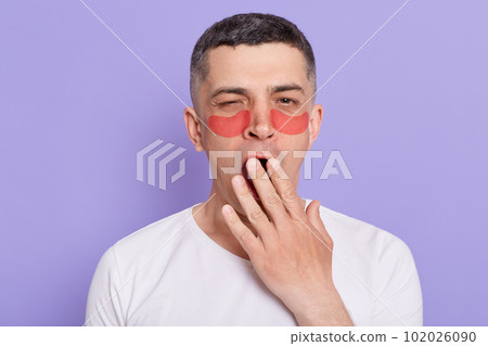 Indoor shot of sleepless man with patches under eyes standing and yawning, covering mouth with palms, feels sleepy, posing isolated on purple background. Indoor shot of sleepless man with patches under eyes standing and yawning, covering mouth with palms, feels sleepy, posing isolated on purple background. 102026090