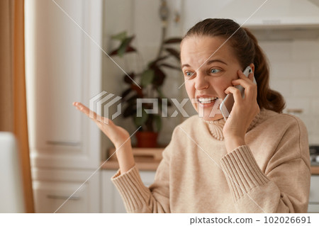 Portrait of angry crazy young adult woman wearing beige sweater sitting in kitchen and talking via mobile phone, spreading hand aside and screaming with hate. Portrait of angry crazy young adult woman wearing beige sweater sitting in kitchen and talking via mobile phone, spreading hand aside and screaming with hate. 102026691