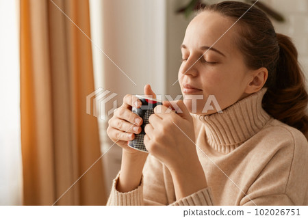 Indoor shot of joyful Caucasian woman with ponytail hairstyle wearing beige casual style sweater posing at home with cup of hot beverage, enjoying morning coffee or tea. 102026751