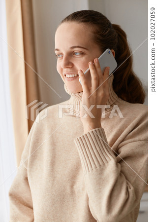 Portrait of optimistic delighted winsome woman with dark hair and ponytail wearing beige jumper talking via cell phone and looking away, expressing pleasant emotions. Portrait of optimistic delighted winsome woman with dark hair and ponytail wearing beige jumper talking via cell phone and looking away, expressing pleasant emotions. 102027059