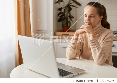 Portrait of positive woman with ponytail hairstyle wearing casual style beige sweater sitting at table in kitchen and looking at laptop screen, holding hands under chin, watching movie or webinar. Portrait of positive woman with ponytail hairstyle wearing casual style beige sweater sitting at table in kitchen and looking at laptop screen, holding hands under chin, watching movie or webinar. 102027086