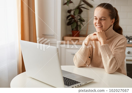 Indoor shot of smiling woman wearing beige sweater sitting in kitchen and having online lesson on laptop, looking at notebook screen with pleasant smile, having video call. 102027087
