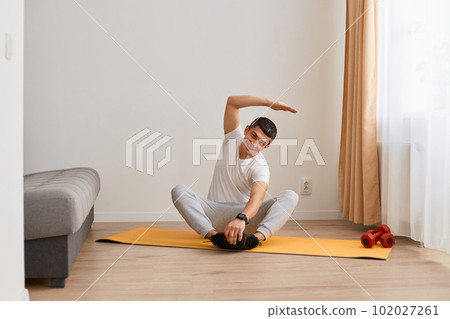 Brunette man wearing sportswear doing sport exercises at home, sitting in lotus position on mat practicing yoga, bending to side with raised hand, doing stretching workouts. 102027261