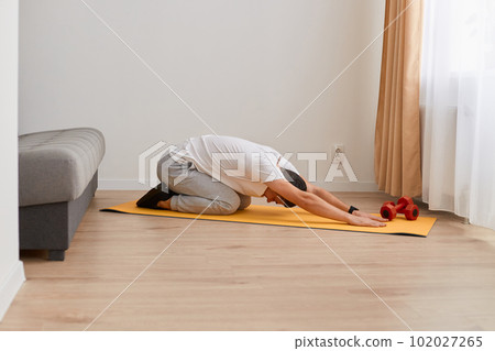 Image of sporty man stretching and practicing yoga at home, young man doing exercises, posing indoor at home on yoga mat, training flexibility, wearing white t shirt and gray pants. 102027265