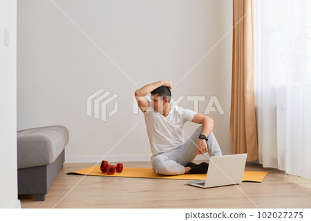 Indoor shot of young adult Caucasian brunette man sniffing his armpit after workout at home on floor on yoga mat, needs antiperpirant, fitness and sport. 102027275