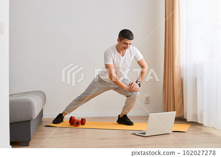 Fit gymnast man with dark hair in sportswear standing on mat and stretching legs, warming up, doing sports flexibility exercises, fitness and yoga workout at home. Fit gymnast man with dark hair in sportswear standing on mat and stretching legs, warming up, doing sports flexibility exercises, fitness and yoga workout at home. 102027278