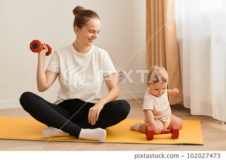 Indoor shot of smiling woman sitting on yoga mat with her baby daughter and training her arms, her biceps and triceps, doing sport exercises and looking after toddler kid. 102027473
