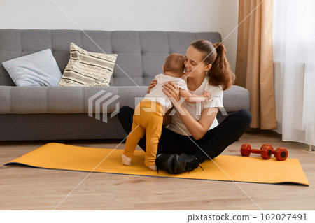 Indoor shot of sporty woman wearing white t shirt and black leggins doing sport exercises at home, hugging her baby daughter while having rest, expressing love. 102027491