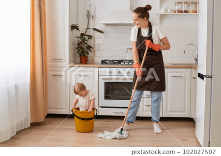 Full length portrait of young adult housewife cleaning house with her little cute charming daughter sitting in bucket, having fun with child while washing floor in kitchen. Full length portrait of young adult housewife cleaning house with her little cute charming daughter sitting in bucket, having fun with child while washing floor in kitchen. 102027507