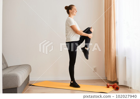 Side view full length of sporty woman wearing white t shirt and black leggins doing sport exercises at home, standing with raised leg, looking ahead, posing near sofa in front of window. 102027940