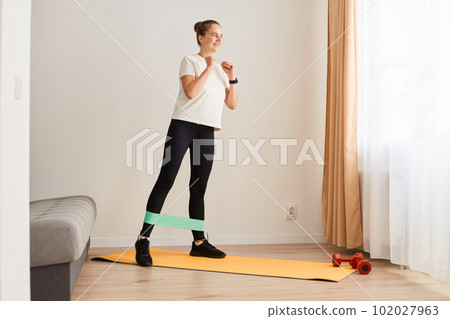 Full and lower body workout. Sporty beautiful woman exercising with elastic rubber band lifting and kicking leg aside standing on yoga mat, Bodyweight training 102027963