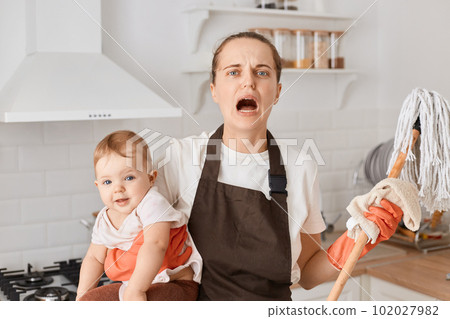 Indoor shot of unhappy despair housewife mother wearing white t shirt and brown apron, holding mop and infant baby in hands, posing in kitchen, screaming, being tired. Indoor shot of unhappy despair housewife mother wearing white t shirt and brown apron, holding mop and infant baby in hands, posing in kitchen, screaming, being tired. 102027982