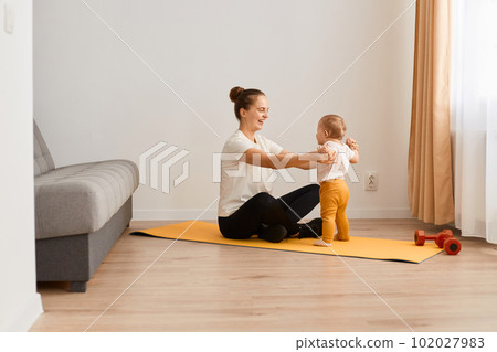Horizontal shot of attractive Caucasian woman wearing white t shirt and black leggins doing sport exercises at home, baby daughter standing near mother, spending time together. Horizontal shot of attractive Caucasian woman wearing white t shirt and black leggins doing sport exercises at home, baby daughter standing near mother, spending time together. 102027983