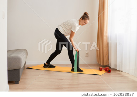 Portrait of sporty woman at home trying to lose weight, standing on rubber stripe training legs, exercising with rubber expander indoor, resistance band for muscle stretching and strength workouts. Portrait of sporty woman at home trying to lose weight, standing on rubber stripe training legs, exercising with rubber expander indoor, resistance band for muscle stretching and strength workouts. 102028057