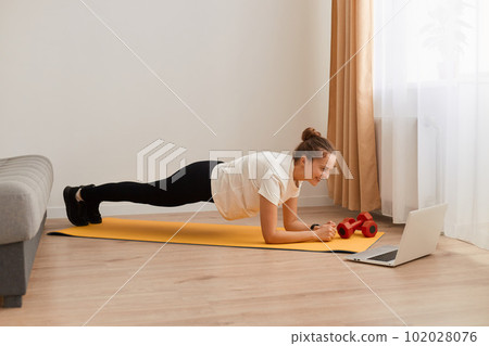 Full length side view portrait of athletic woman wearing white T-shirt and black leggins training her abdominal muscles, standing in plank pose, fitness at home on yoga mat. Full length side view portrait of athletic woman wearing white T-shirt and black leggins training her abdominal muscles, standing in plank pose, fitness at home on yoga mat. 102028076