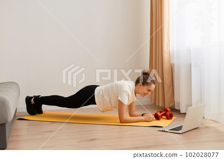 Portrait of attractive woman in sportswear making plank in living room while watching fitness online video on laptop, doing exercises for abs, weight and good physical shape. 102028077