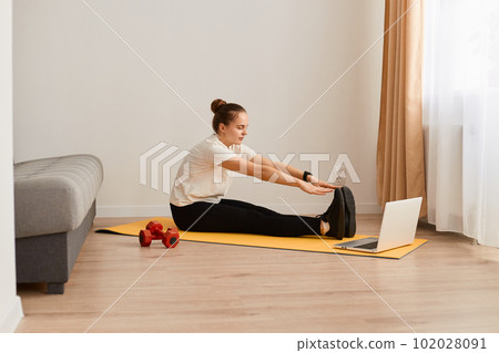 Side view portrait of woman in sportswear watching online video on laptop and repeating after trainer, stretching legs, doing yoga practice, doing exercises for good physical shape. 102028091