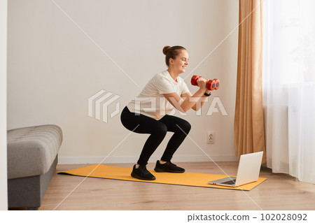 Portrait of athletic woman with hair bun doing squat, lower body sport exercise, keeping balance, warming up and training muscles, holding red dumbbless in hands, wearing white T-shirt and leggins. 102028092