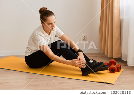 Portrait of ill sick athletic woman wearing white t shirt and black leggins doing sport exercises at home, sitting on mat and keeping hand on painful leg, trauma on workout. 102028118