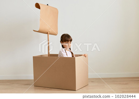 Indoor shot of unhappy little girl playing in handmade ship from cardboard box with sail, expressing negative emotions and sorrow, being boring to play alone. 102028444