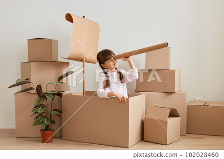 Horizontal shot of pretty little girl making ship from carton box with flag during relocation and playing sailor at new flat, holding big spotting scope and looking far, exploring new islands. 102028460