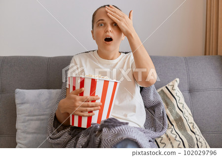 Indoor shot of attractive scared young woman wearing white t shirt sitting on sofa with popcorn and remote control, watching horror film, keeps mouth widely opened, being very impressed. 102029796
