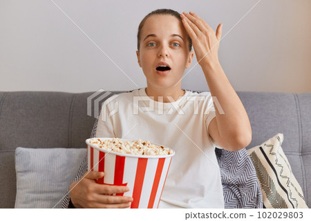 Portrait of astonished young woman wearing white t shirt sitting on sofa with popcorn and remote control, watching tv, impressed by talk show, looking at camera with big eyes. Portrait of astonished young woman wearing white t shirt sitting on sofa with popcorn and remote control, watching tv, impressed by talk show, looking at camera with big eyes. 102029803
