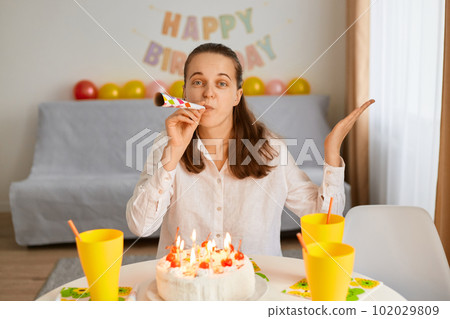 Portrait of happy Caucasian birthday lady blowing party tube while sitting at table in decorated living room, expressing positive emotions, looking at camera and spreading hand. 102029809