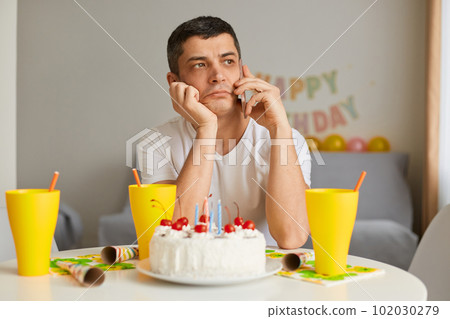 Portrait of sad pensive young man wearing white casual style T-shirt sitting at table with cake, talking smart phone with her friend, celebrating birthday alone, feeling sorrow. 102030279