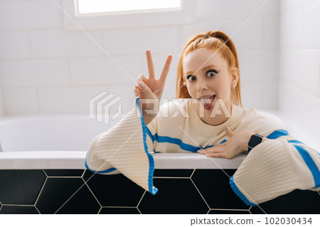 Portrait of fooling funny young woman showing pierced tongue and victory sign sitting in clothes in empty dry bathtub, looking at camera. Attractive female having fun showing horn sign fooling inside Portrait of fooling funny young woman showing pierced tongue and victory sign sitting in clothes in empty dry bathtub, looking at camera. Attractive female having fun showing horn sign fooling inside 102030434