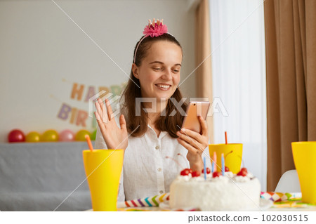 Caucasian positive young woman wearing white shirt celebrating birthday, having video call or livestream, waving hand, saying hello or bye to her friends or relatives. Caucasian positive young woman wearing white shirt celebrating birthday, having video call or livestream, waving hand, saying hello or bye to her friends or relatives. 102030515
