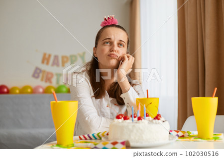 Image of bored attractive young woman wearing white shirt celebrating birthday, sitting alone at table with cake, keeping hand under chin, looking away, dreaming. Image of bored attractive young woman wearing white shirt celebrating birthday, sitting alone at table with cake, keeping hand under chin, looking away, dreaming. 102030516