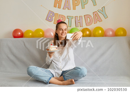 Portrait of young adult happy brown-haired woman smiling sitting on sofa and taking a selfie on a mobile phone camera with a cake with a lit candle, having video call. Portrait of young adult happy brown-haired woman smiling sitting on sofa and taking a selfie on a mobile phone camera with a cake with a lit candle, having video call. 102030530