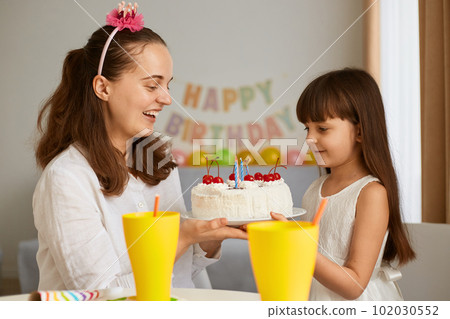 Indoor shot of woman with daughter cerebrating birthday, mother holding cake and congratulating little girl, family posing at home with festive inscription and balloons on background. 102030552
