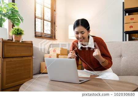 Happy successful small business owner celebrating her highest sales, raising her hands in the with the excitement in a laptop at home 102030680