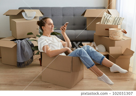 Image of young adult Caucasian woman wearing white t shirt and jeans sitting in a cardboard box and recording voice message, holding smart phone in hands, telling about her relocating. Image of young adult Caucasian woman wearing white t shirt and jeans sitting in a cardboard box and recording voice message, holding smart phone in hands, telling about her relocating. 102031307