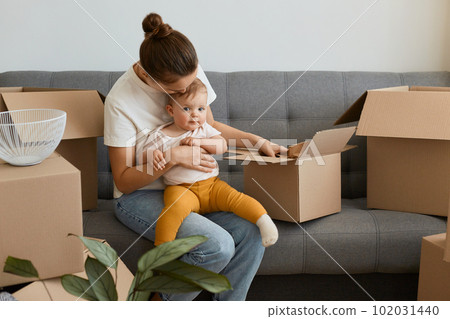Portrait of woman with bun hairstyle wearing white t shirt sitting on sofa and holding her infant baby, unpacking boxes together with her daughter, young family moving into a new apartment. Portrait of woman with bun hairstyle wearing white t shirt sitting on sofa and holding her infant baby, unpacking boxes together with her daughter, young family moving into a new apartment. 102031440