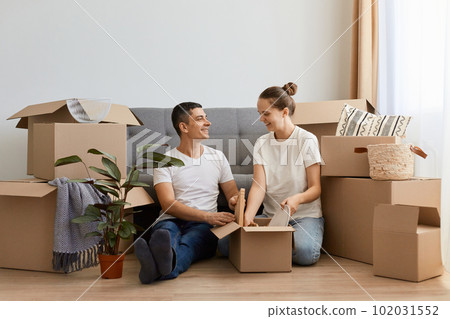 Caucasian family wearing casual attire relocating in new apartment, sitting on the floor near sofa among carton boxes, couple buying a house together, move in new flat, unpacking personal piles. Caucasian family wearing casual attire relocating in new apartment, sitting on the floor near sofa among carton boxes, couple buying a house together, move in new flat, unpacking personal piles. 102031552