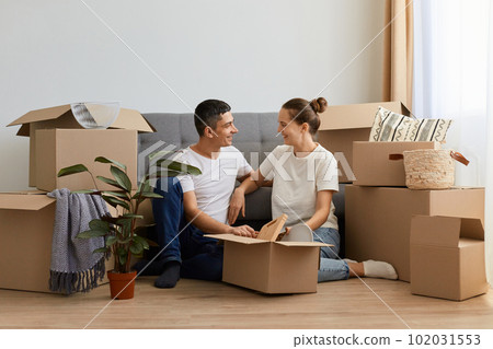 Indoor shot of woman and man wearing white t shirts sitting on floor near sofa being surrounded with boxes with personal belongings, looking at each other with love and happy expression, moving day. 102031553