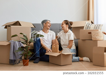 Happy satisfied newlyweds moved in new apartment, sitting on the floor and having fun together, laughing happily, husband with basket on head, relocated in new home. Happy satisfied newlyweds moved in new apartment, sitting on the floor and having fun together, laughing happily, husband with basket on head, relocated in new home. 102031555