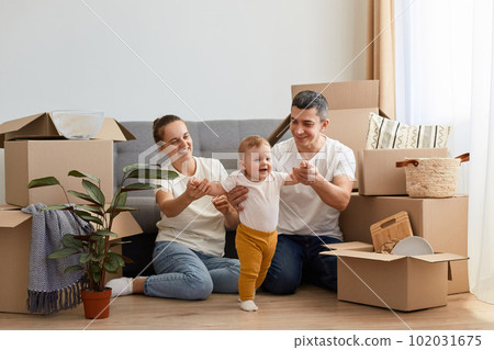 Full length of young couple moving in new apartment, sitting on floor near sofa and playing with their toddler daughter, relocating in a new house, posing among cardboard boxes. 102031675