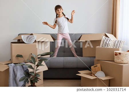 Indoor shot of happy smiling little girl wearing casual style attire standing on sofa and dancing, being happy to move into a new apartment, expressing positive emotions. 102031714